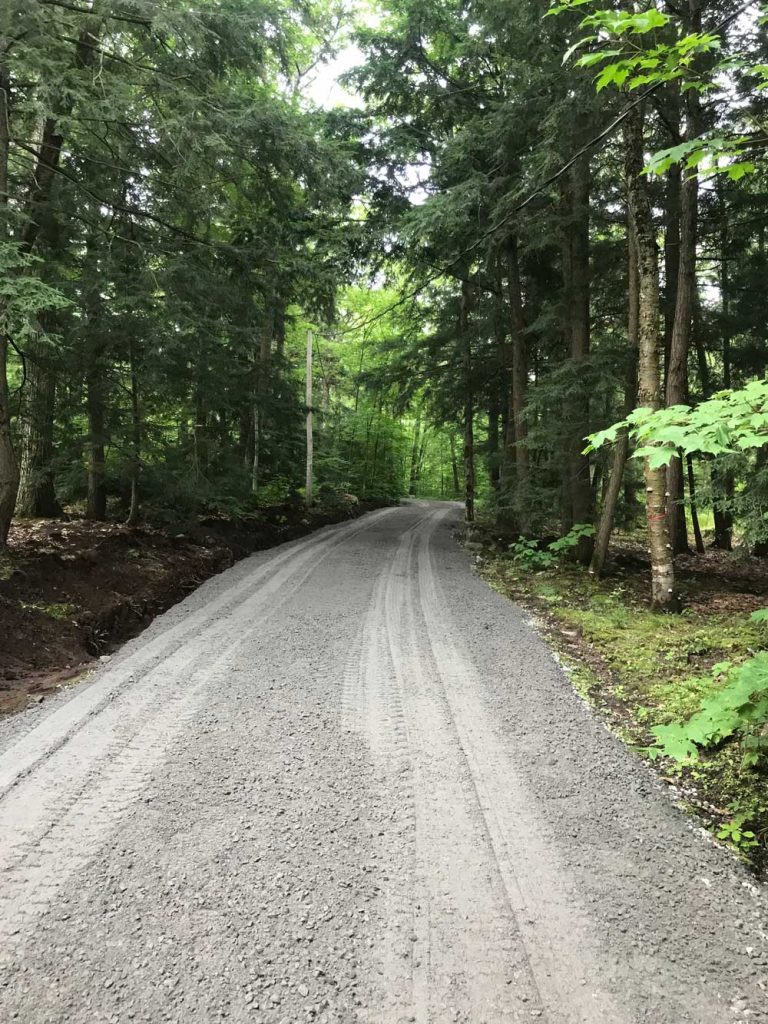 A gravel road runs through a dense forest, surrounded by tall trees with lush green foliage. The road is slightly curved and has tire tracks visible. Sunlight filters through the canopy, casting dappled light and shadows on the ground. The scene is tranquil and scenic. Greenleaf Excavation Muskoka