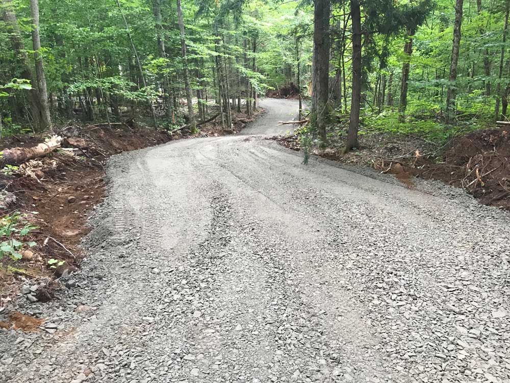 A gravel road curves through a dense forest filled with tall trees and lush green foliage. The road is wide and appears newly constructed with a smooth, even surface. The surrounding area has disturbed earth and debris, likely from the road's recent construction. Greenleaf Excavation Muskoka