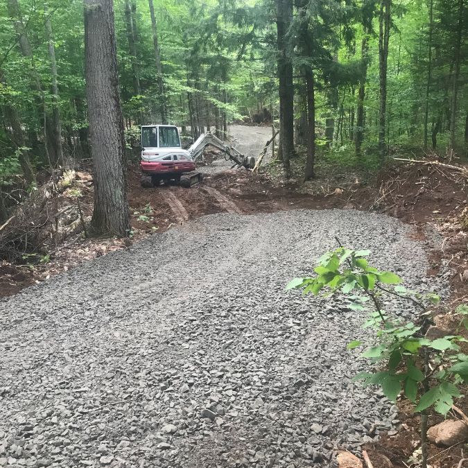 A small excavator works on a gravel path in a forested area. Fresh gravel has been spread over the ground, leading into dense green foliage. The path extends out of view, bordered by a mix of tall trees and underbrush. The scene is bright, indicating daylight under a canopy of leaves. Greenleaf Excavation Muskoka