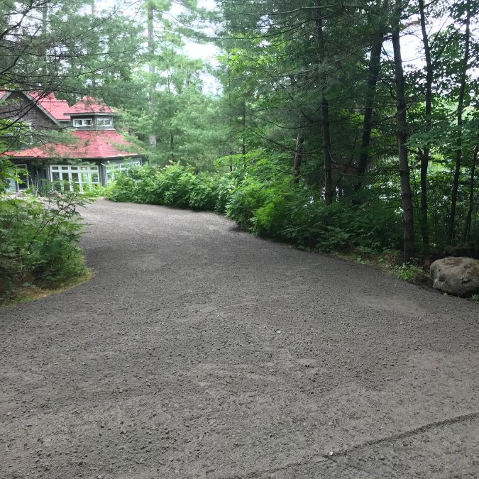 A narrow gravel path winds through a lush green forest with tall trees on each side, leading towards a quaint house with a red roof and large windows. Sunlight filters through the canopy, casting dappled shadows on the path, and dense foliage lines the path's edges. Greenleaf Excavation Muskoka