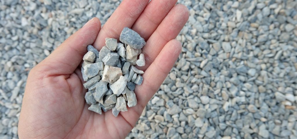 What type of gravel do I need? 6 A hand holds a small pile of grey and white gravel stones. The background is filled with a large amount of similar gravel stones, creating a uniform, textured surface. The stones are irregularly shaped and vary slightly in size. Greenleaf Excavation Muskoka