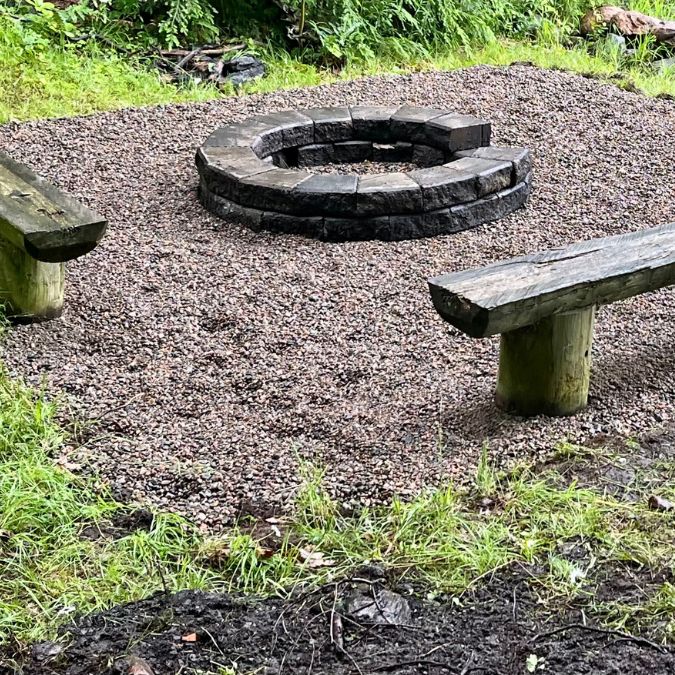 A circular stone fire pit surrounded by gravel is centered in the image. Two rustic wooden benches, created from split logs, are positioned opposite each other on the gravel. Lush green grass and foliage frame the scene, creating a natural and inviting outdoor setting. Greenleaf Excavation Muskoka
