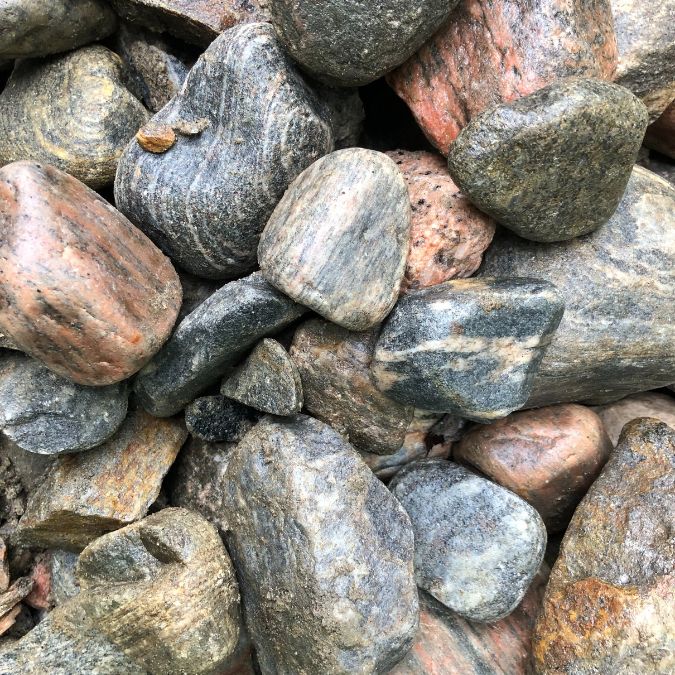 A pile of variously colored smooth rocks and stones. The stones display a range of earthy shades including gray, brown, and hints of orange. They vary in size and shape, with some being rounded and others more irregularly shaped, creating a textured, natural composition. Greenleaf Excavation Muskoka