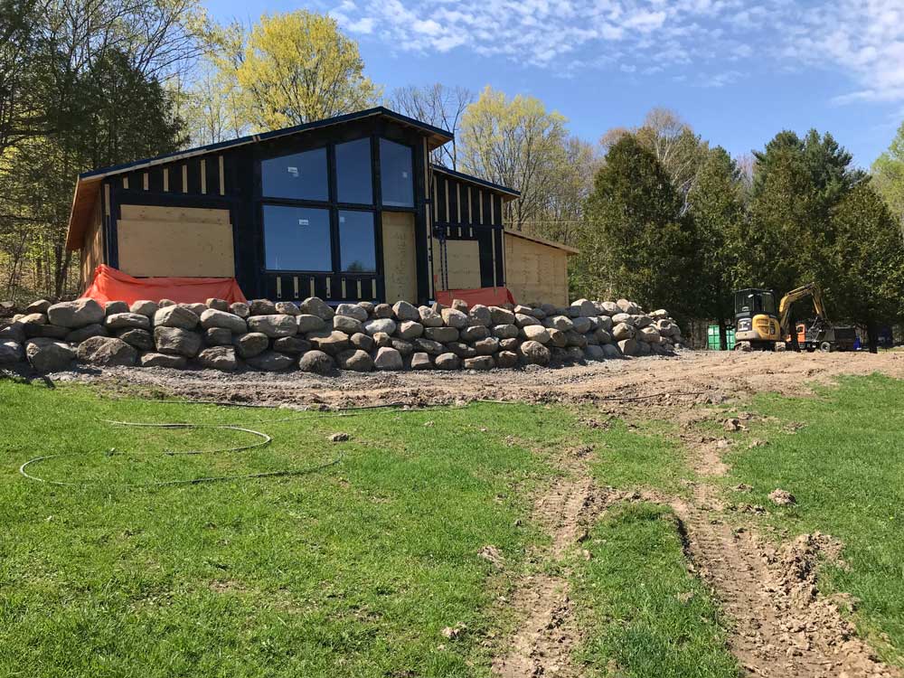 A modern house under construction with large windows, surrounded by a retaining wall of boulders. The house is elevated on a slope with green grass and bare patches in the foreground. Construction equipment is nearby, and trees with spring foliage are in the background. Greenleaf Excavation Muskoka