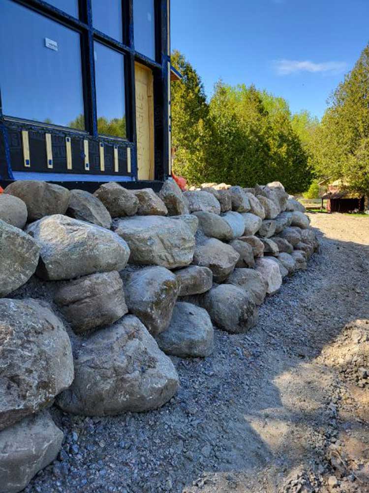 A neatly arranged stone retaining wall built from large, irregularly shaped rocks runs alongside a structure with a black-framed window. The ground is covered with gravel and dirt, while lush green trees and a small red structure are visible in the background under a clear blue sky. Greenleaf Excavation Muskoka