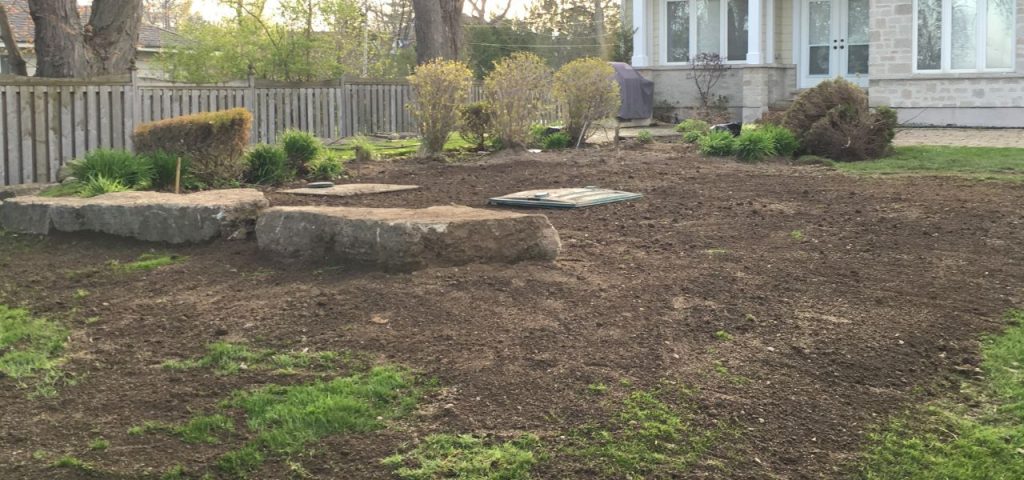 Home and cottage septic system FAQs 4 A garden area with freshly tilled soil surrounded by a wooden fence. Large stone slabs are centrally placed, and small green shrubs are scattered around. In the background, a white house with large windows and a patio is visible, along with a barbecue grill, garden tools, and information on septic systems faqs. Greenleaf Excavation Muskoka