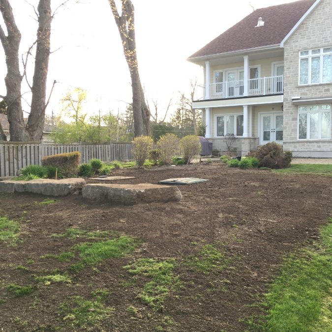A landscaped backyard with freshly tilled soil, a few large rocks placed around the garden, and sparse plants. A beige two-story house with white trim and a balcony is in the background. A wooden fence runs along the left side of the yard, while trees without leaves are visible behind, offering a serene setting perfect for exploring septic systems faqs. Greenleaf Excavation Muskoka