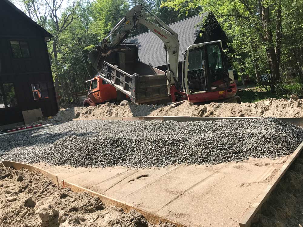 A construction site with an excavator transferring gravel from a pile into a dump truck. The ground is covered with a layer of gravel, and wooden frames are set around the area. Two dark-colored buildings are in the background, surrounded by tall trees and greenery. Greenleaf Excavation Muskoka