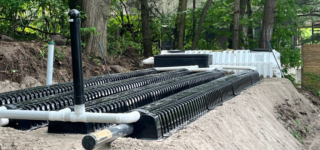 A septic system drain field under construction in a wooded area, featuring rows of black plastic chambers and white PVC pipes on a bed of sand, surrounded by trees and greenery. Greenleaf Excavation Muskoka