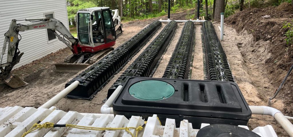 A septic drain field installation in progress shows three long rows of black plastic chambers connected by white pipes near a white building. A small excavator is on the left, and dirt piles and trees are visible in the background. Greenleaf Excavation Muskoka