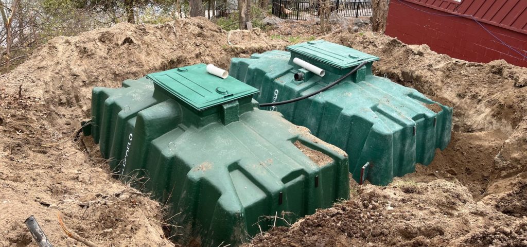 Two large green plastic septic tanks are partially buried in a dirt excavation site, with soil piled around them. Each tank has a rectangular access hatch and white PVC pipes protruding from the lids. Trees and a red building are visible in the background. Greenleaf Excavation Muskoka