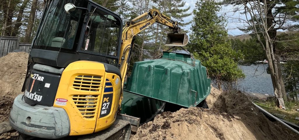 A yellow Komatsu mini excavator lifts a large green tank partially buried in sand, near a forested lakeshore. Trees, a fence, and water are visible in the background, suggesting a construction or installation project by the water. Greenleaf Excavation Muskoka