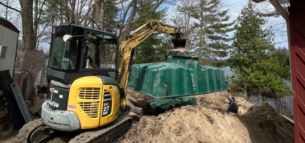 A yellow mini excavator lifts a large green plastic tank over a dirt pit near a lake, surrounded by trees. The tank is labeled 