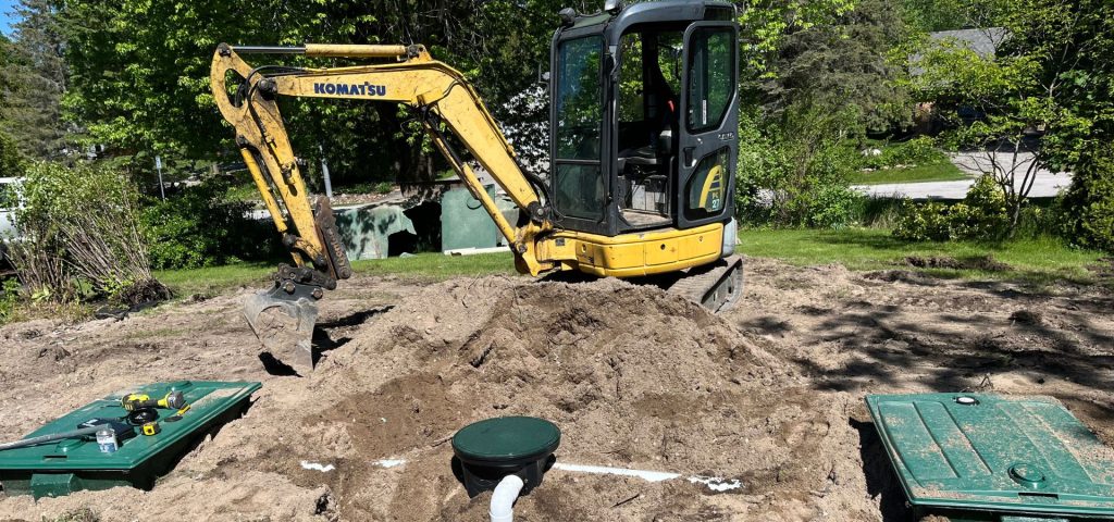 A small yellow Komatsu excavator is parked on a dirt mound in a yard. Nearby are two green septic tanks, one open with tools on top. Trees and a road are visible in the background under bright sunlight. Greenleaf Excavation Muskoka