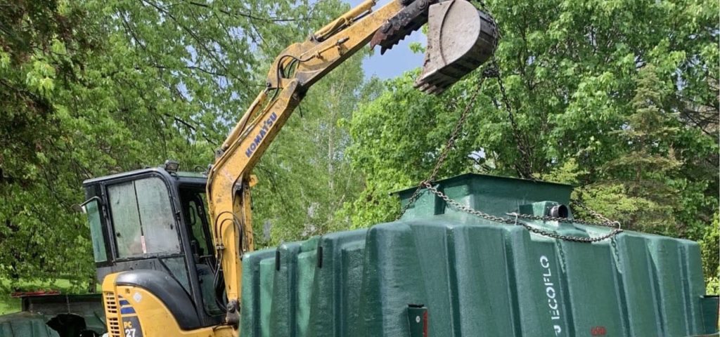 A yellow excavator lifts a large green tank marked 