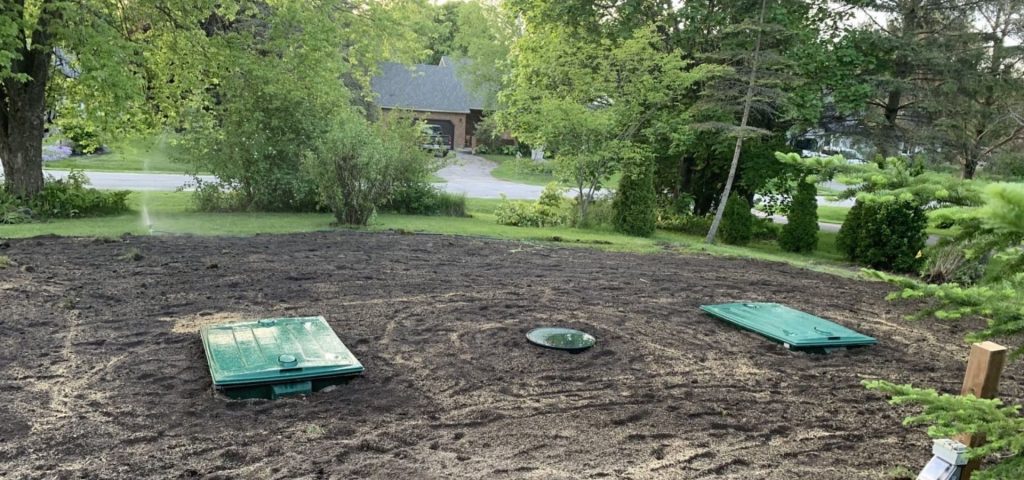 A backyard with two green septic tank covers and a circular access hatch on bare soil. Lush trees and grass surround the area, with a sprinkler watering in the background. A suburban house and driveway are visible beyond the yard. The scene is bright and leafy. Greenleaf Excavation Muskoka