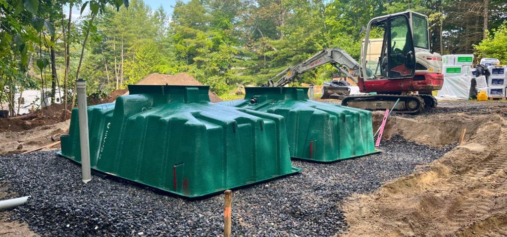 Two large green plastic septic tanks are installed side by side on a bed of black gravel at a construction site. A small excavator is parked nearby, with trees and construction materials visible in the background under daylight. Greenleaf Excavation Muskoka