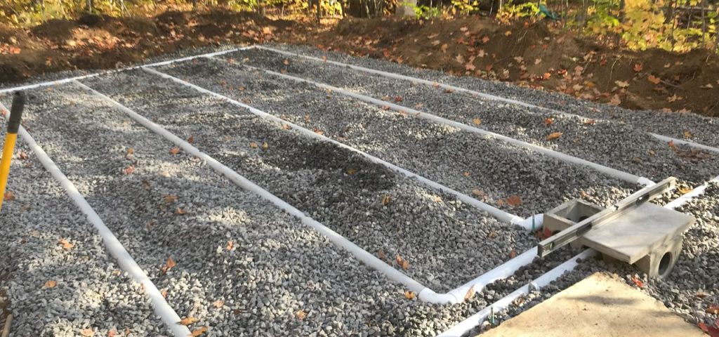 A gravel-covered area with several evenly spaced white PVC pipes arranged in parallel runs. A small rectangular utility box and concrete slab are at one corner. Dirt piles and autumn leaves are visible in the background, suggesting a construction or drainage project. Greenleaf Excavation Muskoka