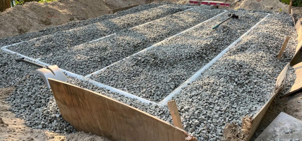 A rectangular construction form filled with gray gravel sits in a dirt area. Wooden boards outline the edges and divide sections. Shovels and leveling tools are placed on top, and piles of dirt surround the form, indicating ongoing construction work. Greenleaf Excavation Muskoka