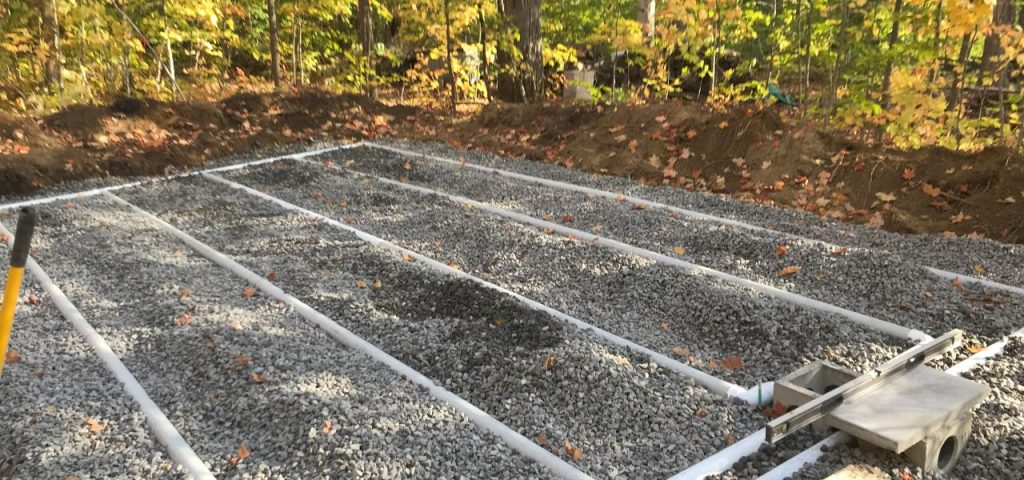 A gravel-covered area is laid out with parallel white PVC pipes, likely part of a septic system drain field installation. In the background, a dirt mound borders a forested area with colorful autumn foliage. A level and shovel are visible at the edge. Greenleaf Excavation Muskoka