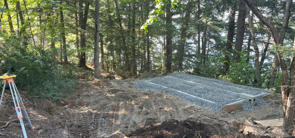 A forested area with sunlight filtering through the trees shows construction work. In the foreground, there are dirt tire tracks and a survey tripod. Midground features a rectangular gravel bed with white pipes, bordered by forest and overlooking a body of water. Greenleaf Excavation Muskoka