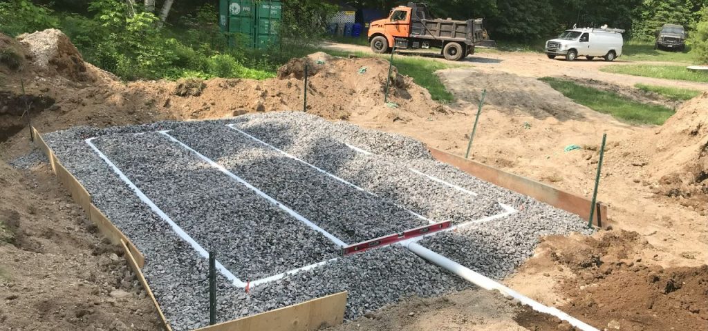 A gravel bed with white PVC pipes arranged in a grid pattern is surrounded by wooden boards and soil piles at a construction site. Trees, a green container, an orange dump truck, a white van, and a black SUV are visible in the background. Greenleaf Excavation Muskoka