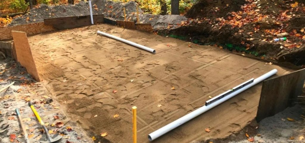 A construction site shows a rectangular, sand-leveled foundation bordered by wooden forms, with two white PVC pipes laid across it. Tools and piles of dirt are nearby, and fallen autumn leaves are scattered around the area. Greenleaf Excavation Muskoka