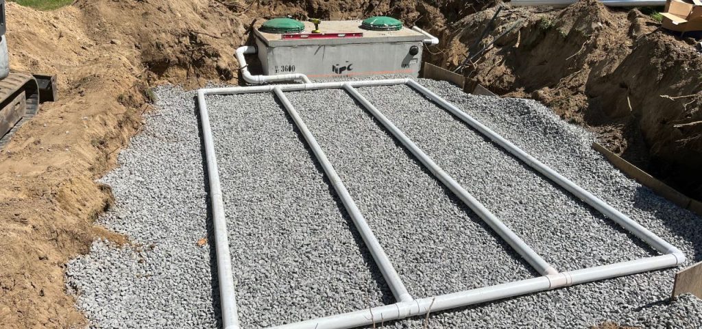 A septic system installation in progress: a rectangular gravel bed with four parallel white PVC pipes is in the foreground, while a concrete septic tank with two green covers and labeled 