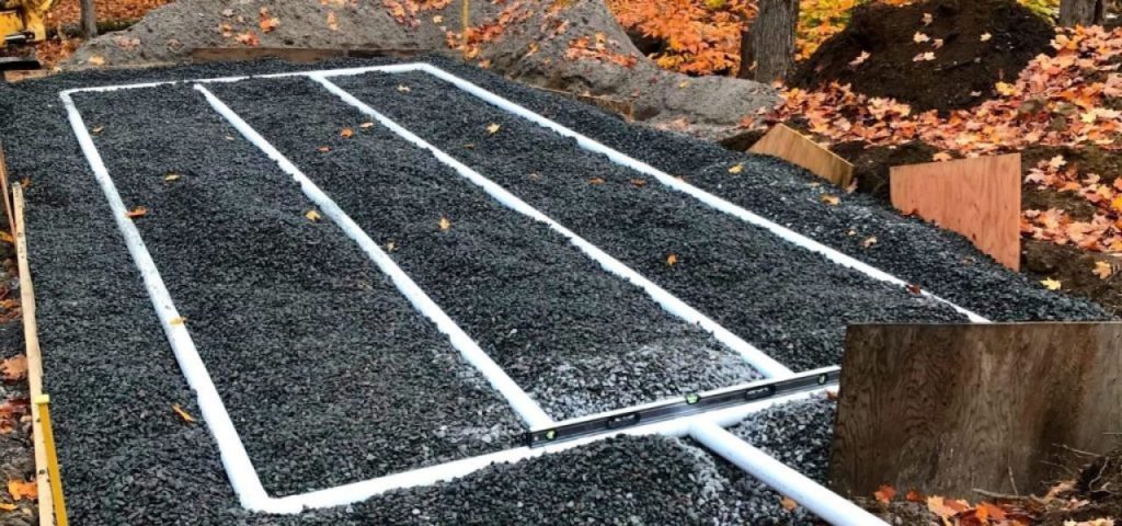 A gravel bed with three white PVC pipes arranged in parallel rows is set outdoors amid fallen autumn leaves. Wooden boards border the area, and soil piles are nearby, indicating a construction site for a drainage or septic system. Greenleaf Excavation Muskoka