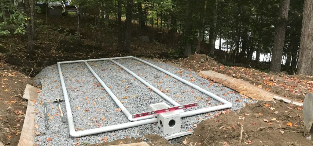 A septic drain field installation in a wooded area, featuring four parallel white PVC pipes laid on a bed of gravel. A spirit level rests on top of the central pipe, and fallen leaves are scattered across the gravel and surrounding soil. Trees and a lake are visible in the background. Greenleaf Excavation Muskoka