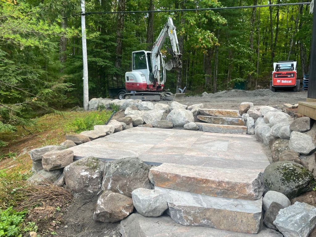 A stone patio with wide stone steps is surrounded by large rocks. Behind it, a white and red mini excavator and red skid-steer sit on gravel near a forest of green trees, suggesting ongoing landscaping or construction work. Greenleaf Excavation Muskoka