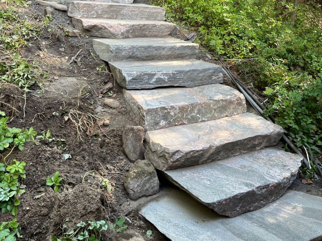 Stone steps made from large, uneven slabs are set into a dirt slope surrounded by green plants and bushes. The stones are rough and irregular, blending with the natural landscape. Some soil and rocks are visible at the edges of the steps. Greenleaf Excavation Muskoka