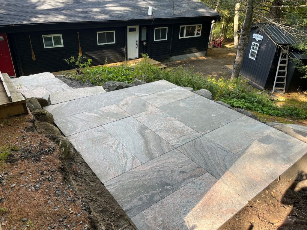 Large, rectangular stone slabs form a newly constructed pathway leading downhill toward two dark wooden buildings surrounded by trees. The path is bordered by rocks, and sunlight filters through the forest, casting dappled shadows on the stones. Greenleaf Excavation Muskoka
